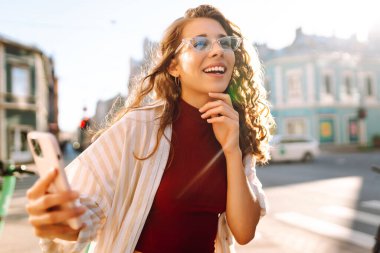 Gülümseyen genç bir kadın eski caddede yürüyor, selfie çekiyor, arkadaşlarla iletişim kuruyor. Ağlar, bloglar, online alışveriş kavramı.