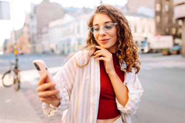 Gülümseyen genç bir kadın eski caddede yürüyor, selfie çekiyor, arkadaşlarla iletişim kuruyor. Ağlar, bloglar, online alışveriş kavramı.
