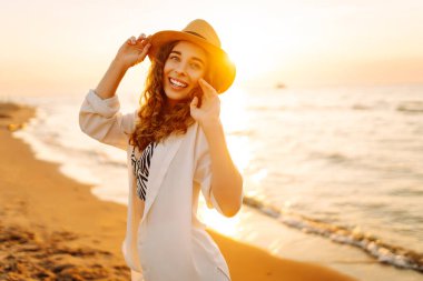 Yazın sahilde mutlu bir kadın moda modası kıyafetleri içinde tasasız ve mutlu, özgürlüğünü hisseden. Gün batımında deniz manzarası ve güzel bir gökyüzü. Seyahat, hafta sonu, rahatlama ve yaşam tarzı konsepti.