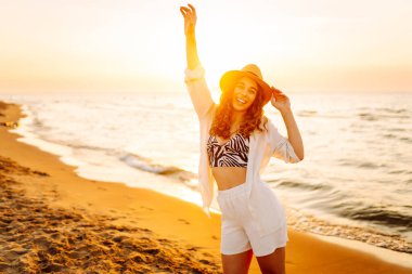 Yazın sahilde mutlu bir kadın moda modası kıyafetleri içinde tasasız ve mutlu, özgürlüğünü hisseden. Gün batımında deniz manzarası ve güzel bir gökyüzü. Seyahat, hafta sonu, rahatlama ve yaşam tarzı konsepti.