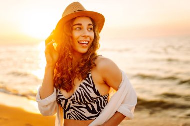 Yazın sahilde mutlu bir kadın moda modası kıyafetleri içinde tasasız ve mutlu, özgürlüğünü hisseden. Gün batımında deniz manzarası ve güzel bir gökyüzü. Seyahat, hafta sonu, rahatlama ve yaşam tarzı konsepti.