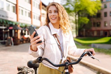 Bisikletle yürürken cep telefonu kullanan genç bir kadın güzel bir mimariye bakıyor. Selfie zamanı. İnsanlar, boş zaman, blog ve yaşam tarzı.