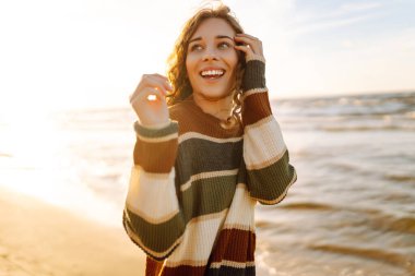 Şık bir kadın serin bir günde deniz ya da okyanus sahillerinde günbatımında yürür. Harika bir açık hava manzarası. Seyahat, hafta sonu, rahatlama ve yaşam tarzı konsepti.