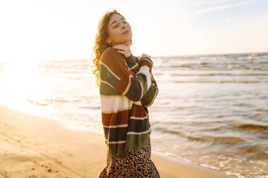 Şık bir kadın serin bir günde deniz ya da okyanus sahillerinde günbatımında yürür. Harika bir açık hava manzarası. Seyahat, hafta sonu, rahatlama ve yaşam tarzı konsepti.