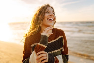 Şık bir kadın serin bir günde deniz ya da okyanus sahillerinde günbatımında yürür. Harika bir açık hava manzarası. Seyahat, hafta sonu, rahatlama ve yaşam tarzı konsepti.