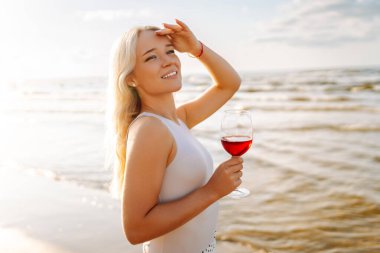 Gün batımında zarif beyaz elbiseler içinde bir bardak şarapla güzel bir manken. Seyahat, hafta sonu, rahatlama ve yaşam tarzı konsepti.