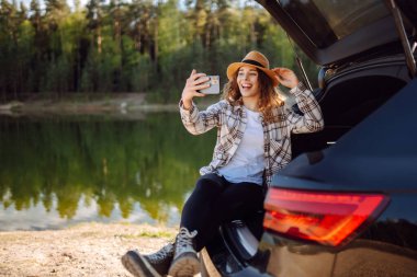 Araba yolculuğu ile selfie. Genç kadın arabanın bagajında dinleniyor ve selfie çekiyor. Seyahat, hafta sonu, rahatlama ve yaşam tarzı konsepti.