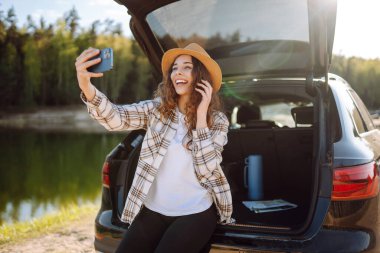Araba yolculuğu ile selfie. Genç kadın arabanın bagajında dinleniyor ve selfie çekiyor. Seyahat, hafta sonu, rahatlama ve yaşam tarzı konsepti.