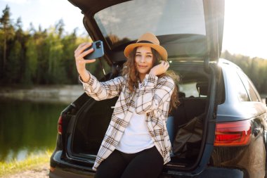 Araba yolculuğu ile selfie. Genç kadın arabanın bagajında dinleniyor ve selfie çekiyor. Seyahat, hafta sonu, rahatlama ve yaşam tarzı konsepti.