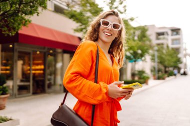 Sokakta dolaşan genç bir kadın akıllı telefon kullanıyor, arkadaşlarıyla iletişim kuruyor. Ağlar, bloglar, online alışveriş kavramı.