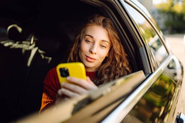Arabada akıllı telefon kullanan mutlu genç bir kadın. Teknoloji kavramı, blog yazma, arabayla seyahat, iş.