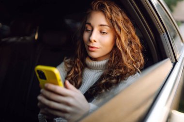 Arabada akıllı telefon kullanan mutlu genç bir kadın. Teknoloji kavramı, blog yazma, arabayla seyahat, iş.