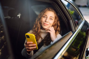 Arabada akıllı telefon kullanan mutlu genç bir kadın. Teknoloji kavramı, blog yazma, arabayla seyahat, iş.
