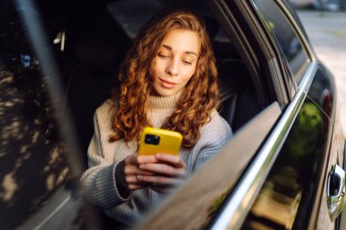 Arabada akıllı telefon kullanan mutlu genç bir kadın. Teknoloji kavramı, blog yazma, arabayla seyahat, iş.