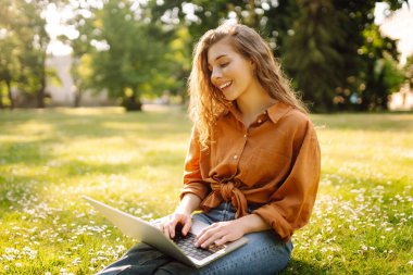 Çimlerin üzerinde oturan genç bir kadın şehir parkındaki bilgisayarla dışarıda çimlerin üzerinde çalışıyor. Mobil Ofis. Serbest ticaret kavramı