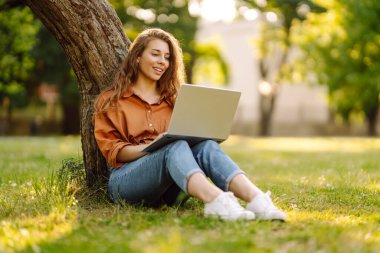 Çimlerin üzerinde oturan genç bir kadın şehir parkındaki bilgisayarla dışarıda çimlerin üzerinde çalışıyor. Mobil Ofis. Serbest ticaret kavramı
