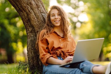 Çimlerin üzerinde oturan genç bir kadın şehir parkındaki bilgisayarla dışarıda çimlerin üzerinde çalışıyor. Mobil Ofis. Serbest ticaret kavramı