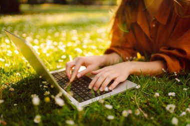 Çimlerin üzerinde oturan genç bir kadın şehir parkındaki bilgisayarla dışarıda çimlerin üzerinde çalışıyor. Mobil Ofis. Serbest ticaret kavramı