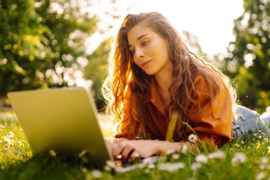 Çimlerin üzerinde oturan genç bir kadın şehir parkındaki bilgisayarla dışarıda çimlerin üzerinde çalışıyor. Mobil Ofis. Serbest ticaret kavramı