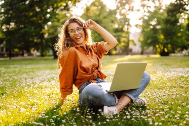 Çimlerin üzerinde oturan genç bir kadın şehir parkındaki bilgisayarla dışarıda çimlerin üzerinde çalışıyor. Mobil Ofis. Serbest ticaret kavramı