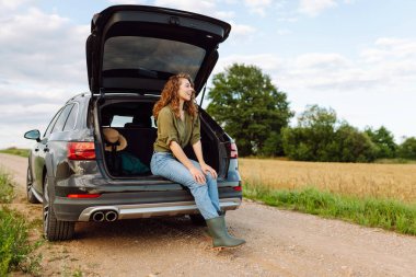 Günbatımının tadını çıkaran genç bir kadın yalnız başına yolculuk yaparken arabasının bagajında oturuyor. Arabayla yolculuk. Yaşam tarzı, seyahat, turizm, doğa konsepti.