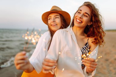 Bengal ışıkları olan neşeli genç kadınlar göl kıyısında dans ediyorlar. Birlikte gülüyorlar. Arka planda güzel bir günbatımı var. Yaz tatili, parti, tatil, dinlenme.