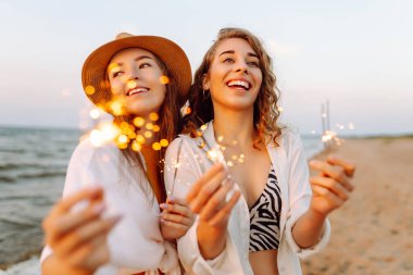 Bengal ışıkları olan neşeli genç kadınlar göl kıyısında dans ediyorlar. Birlikte gülüyorlar. Arka planda güzel bir günbatımı var. Yaz tatili, parti, tatil, dinlenme.