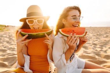 Yazın kumsalda tatilde olan iki mutlu kadın arka planda deniz olan bir karpuz yiyor, gün batımında kumların üzerinde oturuyorlar. Boş zaman ve arkadaşlık kavramı