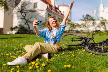 Genç bir kadın parkta yeşil çimlerde oturur ve selfie çeker. Doğada bisiklet sürmek. Seyahat, hafta sonu, rahatlama ve yaşam tarzı konsepti.