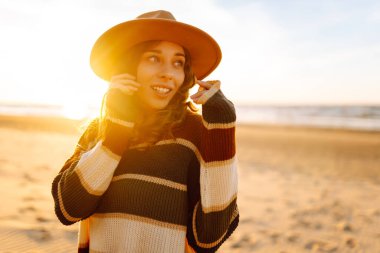 Şık bir kadın gün batımında deniz ya da okyanus sahillerinde gün batımında yürür. Soğuk güneşli ve rüzgarlı bir gün. Seyahat, hafta sonu, rahatlama ve yaşam tarzı konsepti.