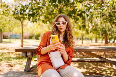 Genç bir kadın parkta bankta tek başına oturuyor, cep telefonu kullanıyor, ekrana bakıyor, mesaj yazıyor, uygulama sohbeti yapıyor. Seyahat, blog, hafta sonu, rahatlama ve yaşam tarzı konsepti.