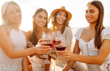 Gülümseyen genç kadınlar kadeh tokuşturup birlikte vakit geçiriyorlar. Kumsalda kızlar partisi. Piknik, tatil, dinlenme, tatil.