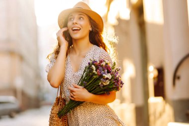 Hasır şapkalı ve elinde sokak şehrinde poz veren kır çiçekleri olan mutlu bir kadın. Moda tarzı.