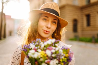 Hasır şapkalı ve elinde sokak şehrinde poz veren kır çiçekleri olan mutlu bir kadın. Moda tarzı.