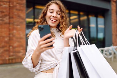 Gülümseyen genç bayan müşteriler ellerinde alışveriş çantaları ve akıllı telefonlar tutuyorlardı. İndirimli ecommerce uygulaması. Tüketim, Kara Cuma. İnternetten alışveriş.