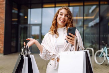 Gülümseyen genç bayan müşteriler ellerinde alışveriş çantaları ve akıllı telefonlar tutuyorlardı. İndirimli ecommerce uygulaması. Tüketim, Kara Cuma. İnternetten alışveriş.