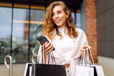 Gülümseyen genç bayan müşteriler ellerinde alışveriş çantaları ve akıllı telefonlar tutuyorlardı. İndirimli ecommerce uygulaması. Tüketim, Kara Cuma. İnternetten alışveriş.