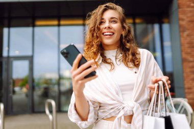 Gülümseyen genç bayan müşteriler ellerinde alışveriş çantaları ve akıllı telefonlar tutuyorlardı. İndirimli ecommerce uygulaması. Tüketim, Kara Cuma. İnternetten alışveriş.