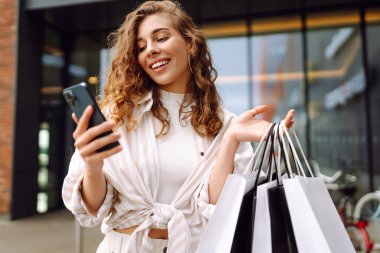 Gülümseyen genç bayan müşteriler ellerinde alışveriş çantaları ve akıllı telefonlar tutuyorlardı. İndirimli ecommerce uygulaması. Tüketim, Kara Cuma. İnternetten alışveriş.