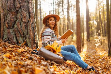Güz parkında oturan ve kitap okuyan güzel gülümseyen genç bayan öğrenci sarı yapraklar dökülüyor. Doğadan zevk alma kavramı.