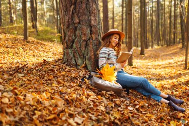Güz parkında oturan ve kitap okuyan güzel gülümseyen genç bayan öğrenci sarı yapraklar dökülüyor. Doğadan zevk alma kavramı.