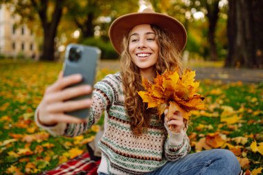 Şapkalı ve örgü süveterli şık bir kadın parkta portakal yapraklı akıllı telefonda selfie çekerken anı yakalıyor. Seyahat blogu. Macera, tatil konsepti.