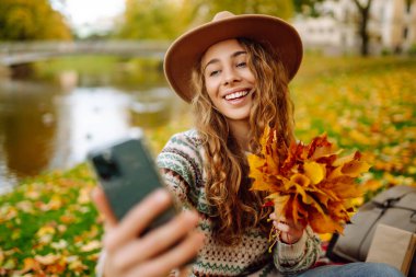Şapkalı ve örgü süveterli şık bir kadın parkta portakal yapraklı akıllı telefonda selfie çekerken anı yakalıyor. Seyahat blogu. Macera, tatil konsepti.