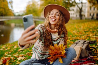 Şapkalı ve örgü süveterli şık bir kadın parkta portakal yapraklı akıllı telefonda selfie çekerken anı yakalıyor. Seyahat blogu. Macera, tatil konsepti.