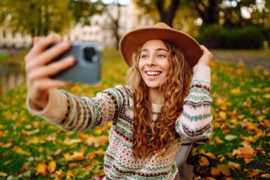 Şapkalı ve örgü süveterli şık bir kadın parkta portakal yapraklı akıllı telefonda selfie çekerken anı yakalıyor. Seyahat blogu. Macera, tatil konsepti.
