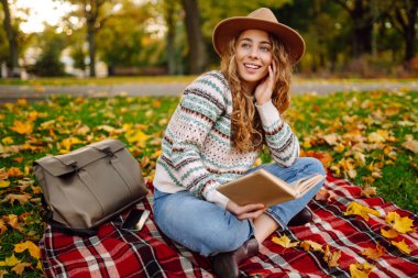 Autumn Park 'ta kitap okuyan genç bir kadın. Sonbahar yürüyüşü kavramı, rahatlama, doğa, piknik.
