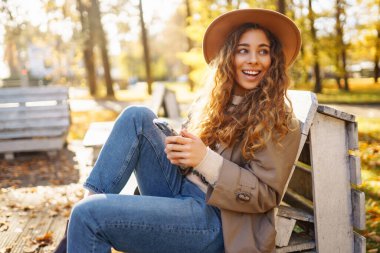 Parktaki sonbahar ağaçlarının arka planında oturan güzel kadın bir bankta oturup akıllı telefondan bir şeye bakıyor. Tatil kavramı, blog yazma, teknoloji, hafta sonu.