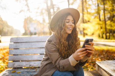 Parktaki sonbahar ağaçlarının arka planında oturan güzel kadın bir bankta oturup akıllı telefondan bir şeye bakıyor. Tatil kavramı, blog yazma, teknoloji, hafta sonu.