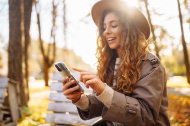 Parktaki sonbahar ağaçlarının arka planında oturan güzel kadın bir bankta oturup akıllı telefondan bir şeye bakıyor. Tatil kavramı, blog yazma, teknoloji, hafta sonu.