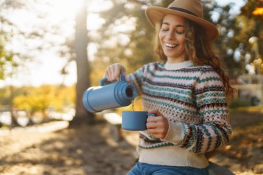 Sonbahar parkında termostan sıcak su içen bir kadın. Tatil, doğada hafta sonu.